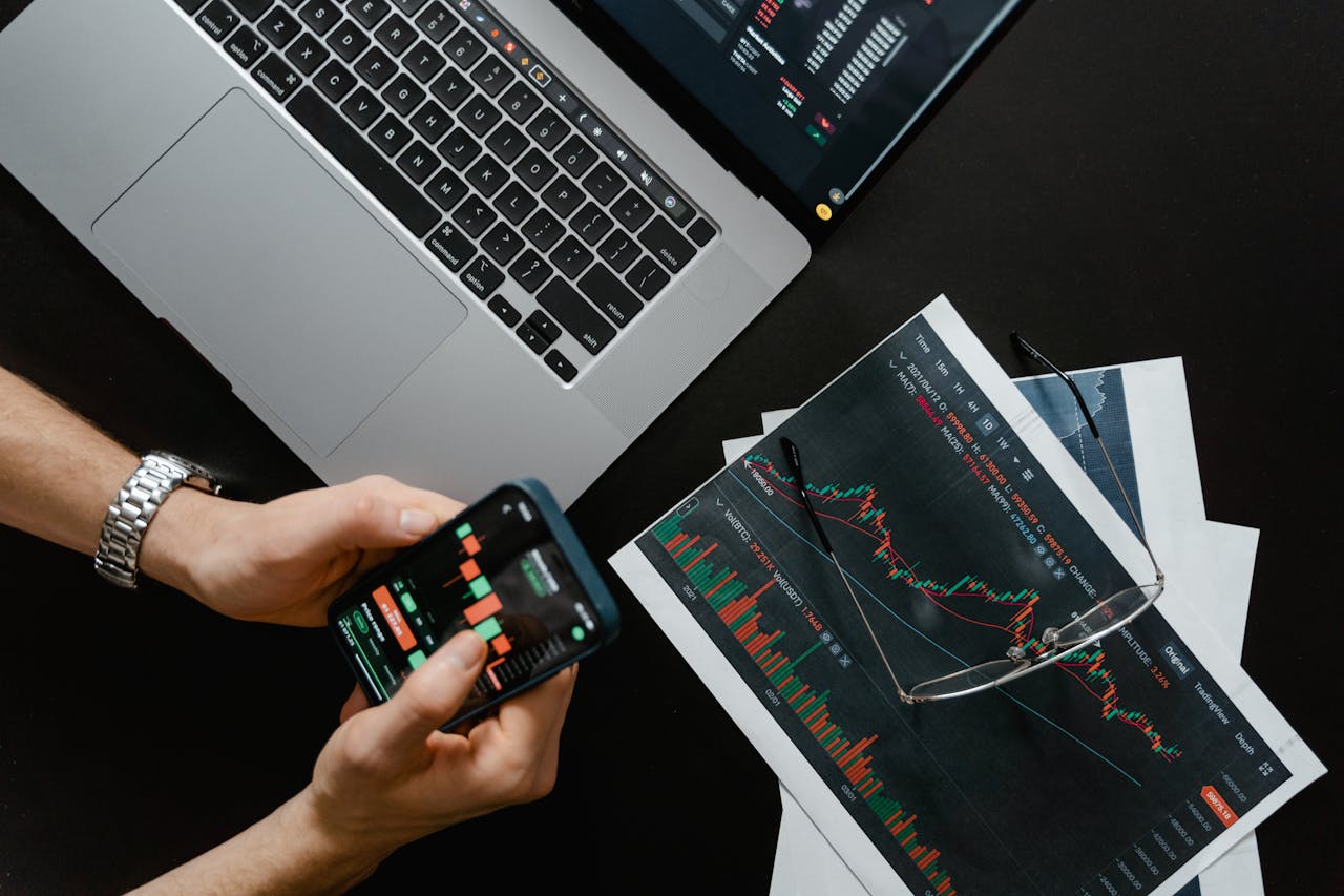 hero-img Hands using smartphone beside laptop with stock charts, showcasing digital trading.
